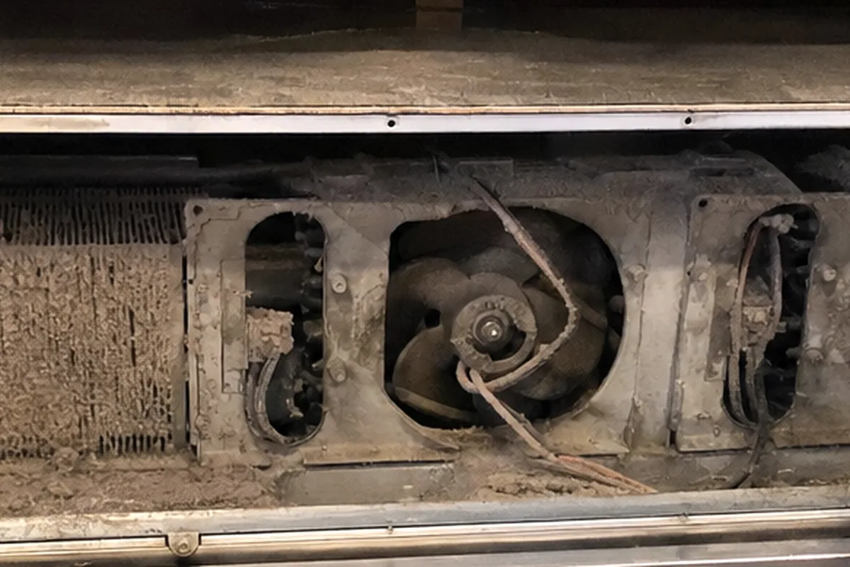 A row of squirrel-cage fans and a finned coil packed with grey lint, seen behind a removed stainless panel